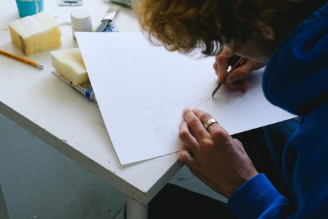 High angle of crop unrecognizable man in casual clothes sitting at table while cutting sketch on paper with utility knife near paints and sponge with pencil in light workshop